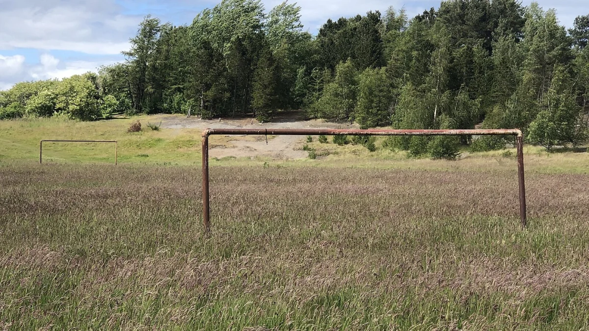Teren de fotbal năpădit de iarbă înaltă, cu o poartă metalică ruginită și pădure în fundal (imagine ilustrativă din Marea Britanie, nu este Baza Triunghi)