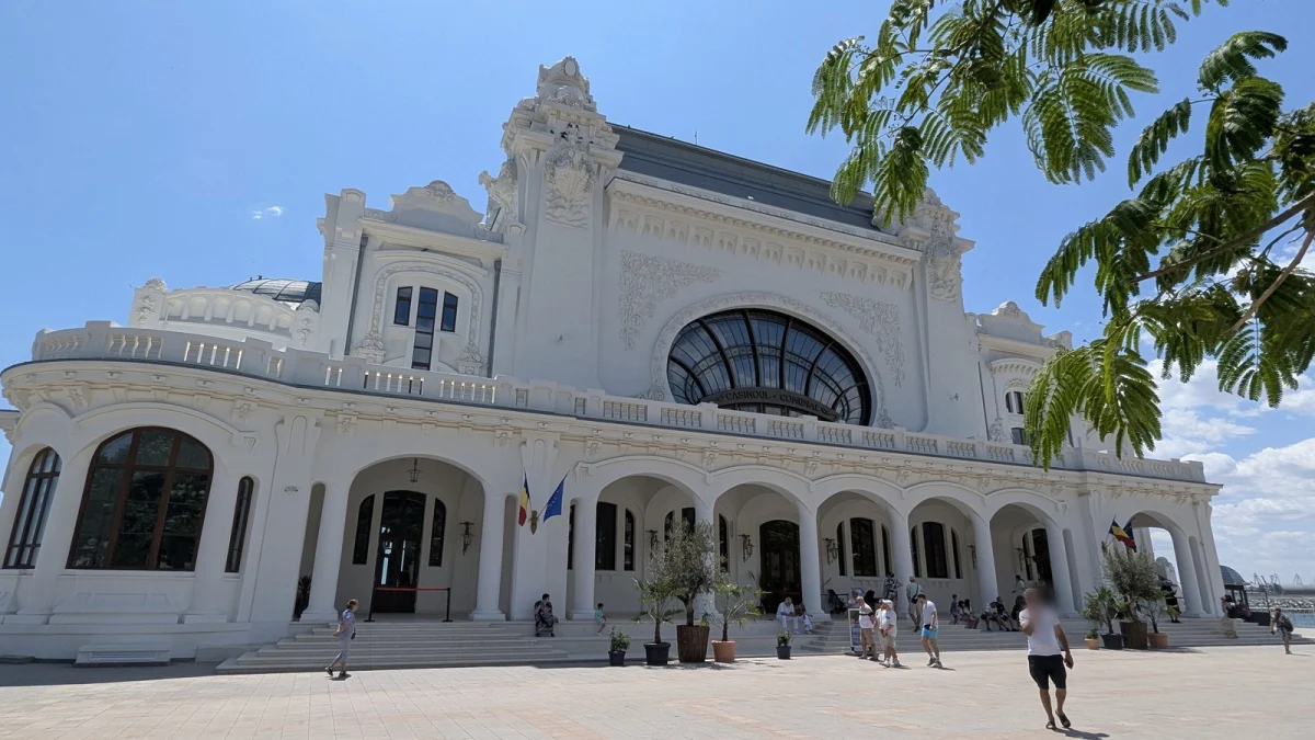 Fațada albă a Cazinoului din Constanța după restaurare, cu arcade, steaguri și trecători pe esplanadă, într-o zi senină de vară (fotografie din 2025)