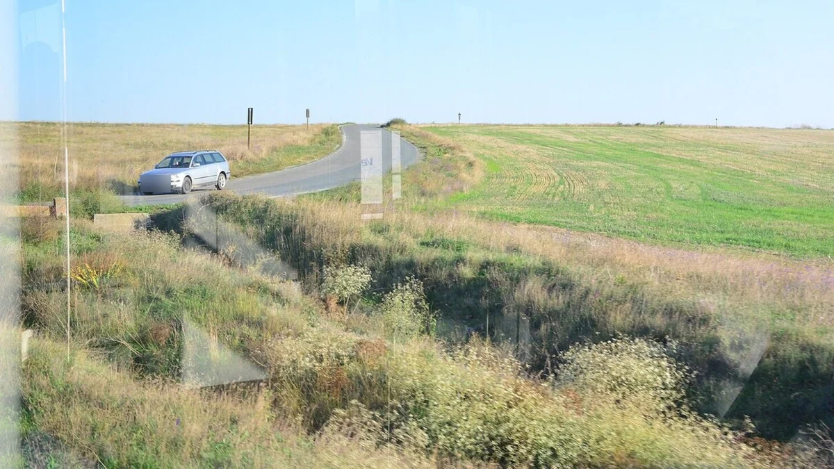 Drumul județean DJ 222 la sud de Târgușor, printre câmpuri, cu un autoturism în curbă, fotografiat prin geamul unui tren (fotografie de arhivă)