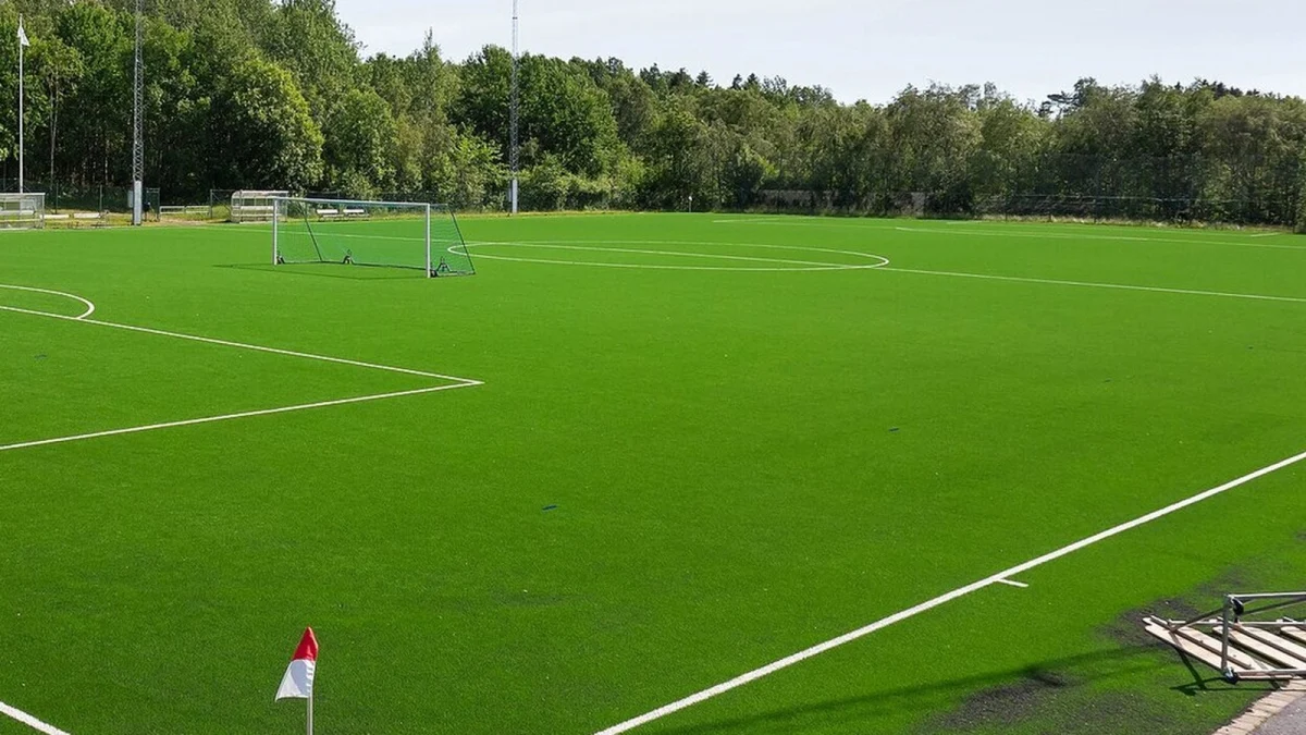 Colț de teren de fotbal cu gazon artificial, steguleț de corner și poartă în fundal, într-o zi senină (imagine ilustrativă, nu este din România)
