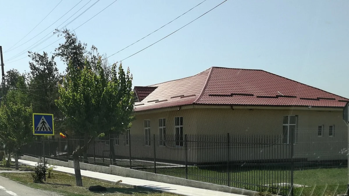 Clădire cu acoperiș roșu din comuna Comana, județul Constanța, în spatele unui gard, cu un indicator de trecere de pietoni (fotografie de arhivă, 2017)